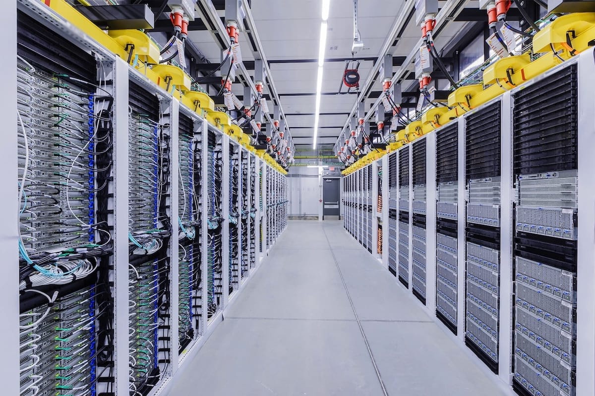A data center aisle: two rows of server racks packed with cabling, tied together by yellow cable trays overhead.